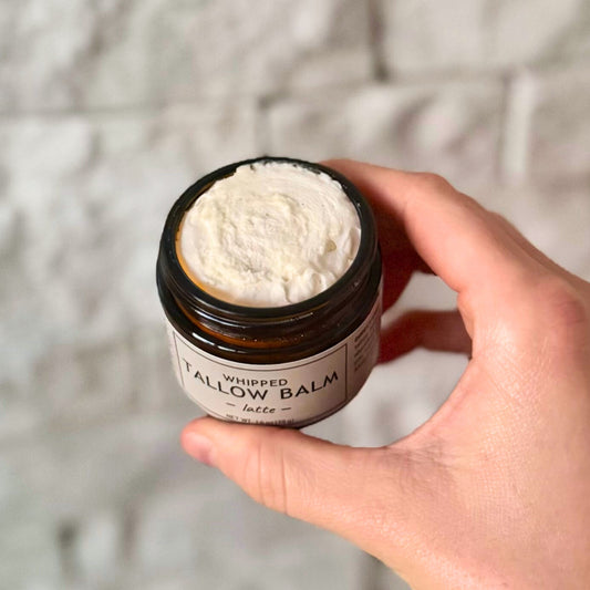 Hand holding a jar of whipped balm labeled 'Tallow Balm' against a neutral background